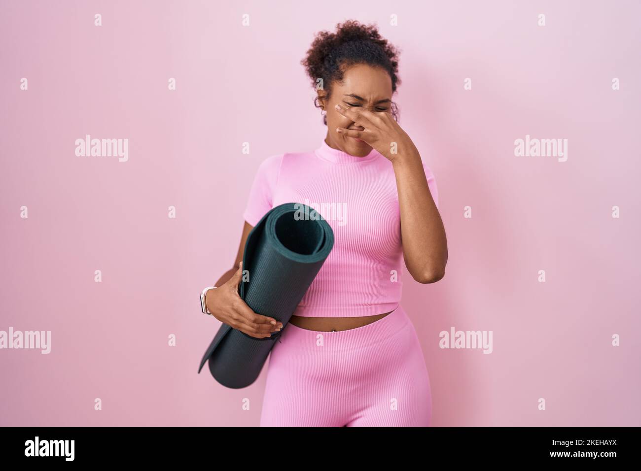 Young hispanic woman with curly hair holding yoga mat over pink