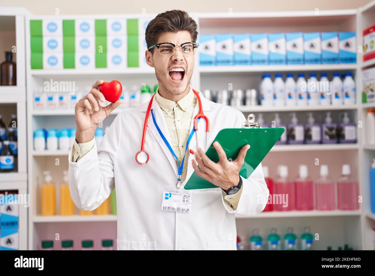 Young hispanic man working at pharmacy drugstore holding heart angry ...