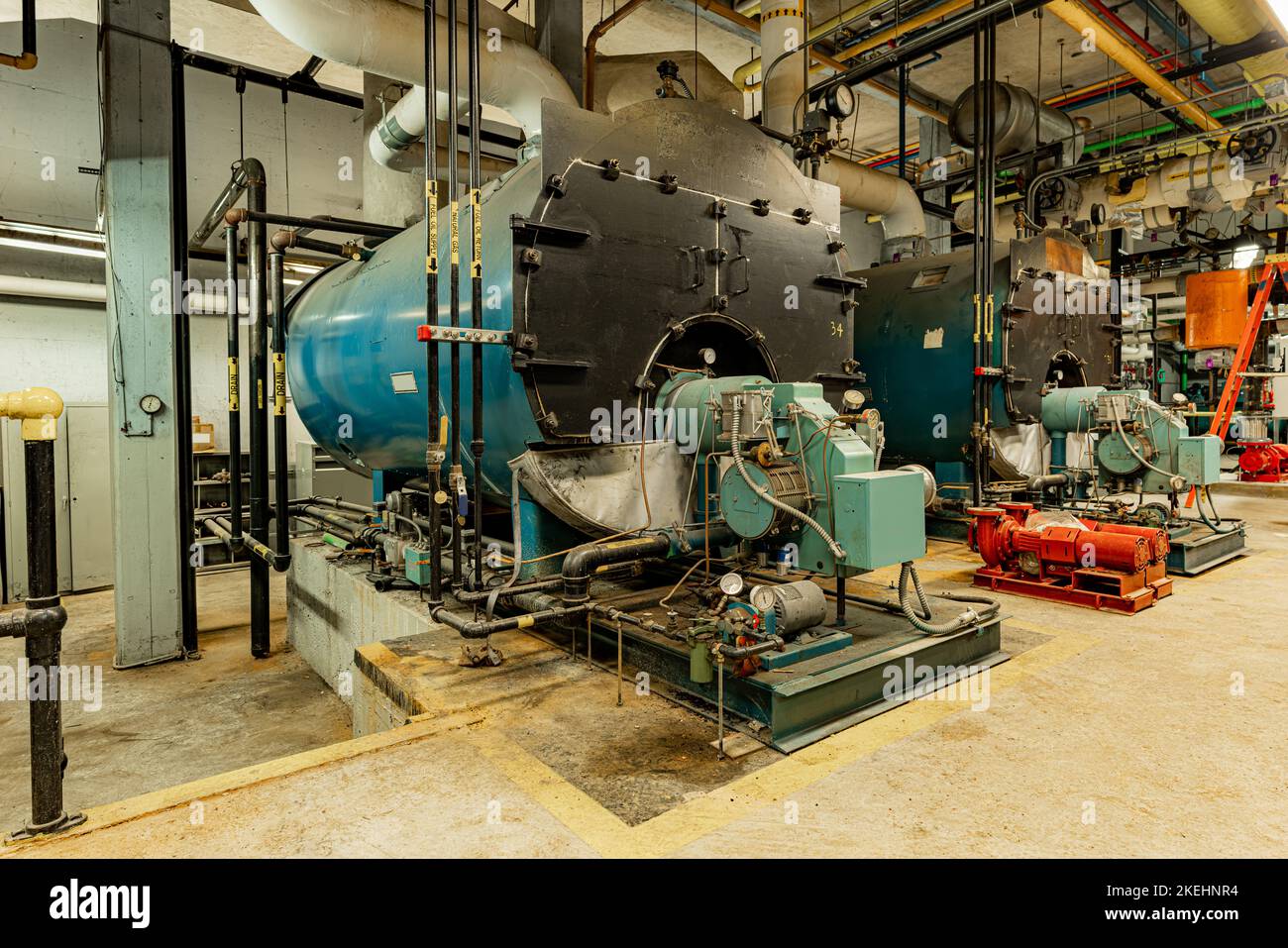 The interior of an old and well used boiler room with two large boilers, many pipes, valves and other equipment. Stock Photo