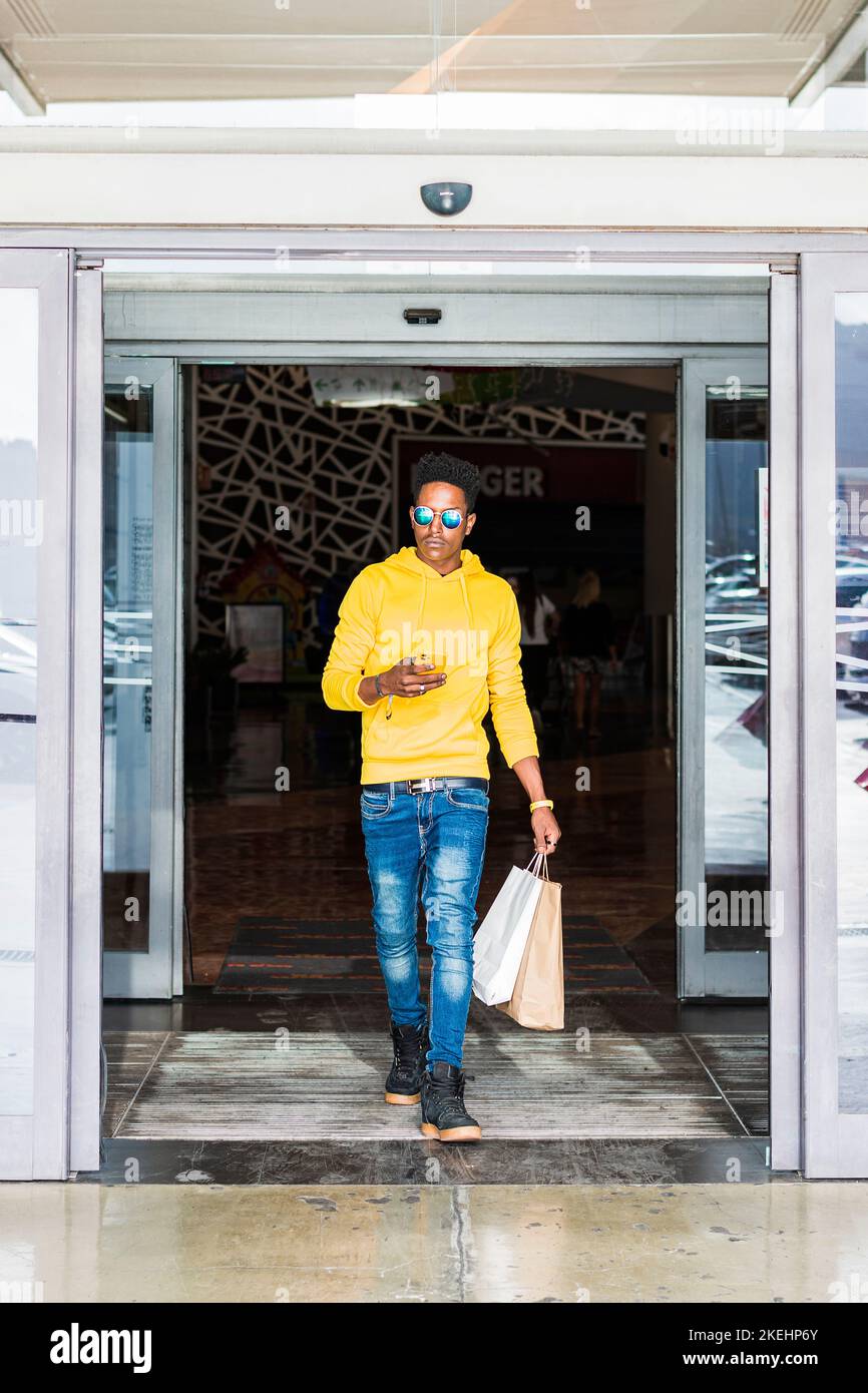A young black adult male wearing a yellow sweatshirt and blue pants is carrying some shopping bags and is leaving a shopping mall while holding his ce Stock Photo