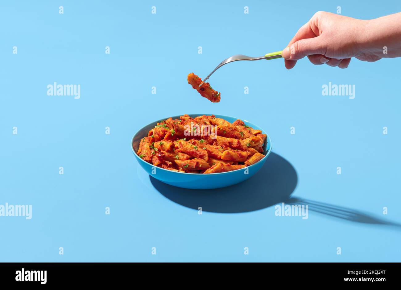 Woman's hand taking a pasta tube from a bowl with a fork. Eating pasta penne alla arrabbiata. Spicy pasta dish in a blue bowl minimalist on a colorful Stock Photo