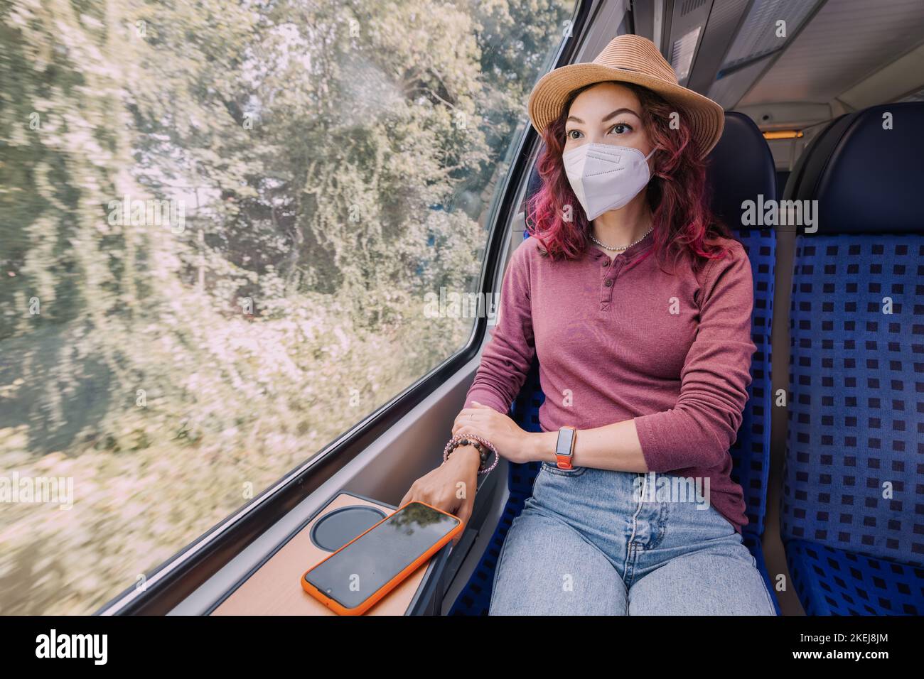 A girl in a protective medical mask rides a high-speed intercity train and looks out the window Stock Photo