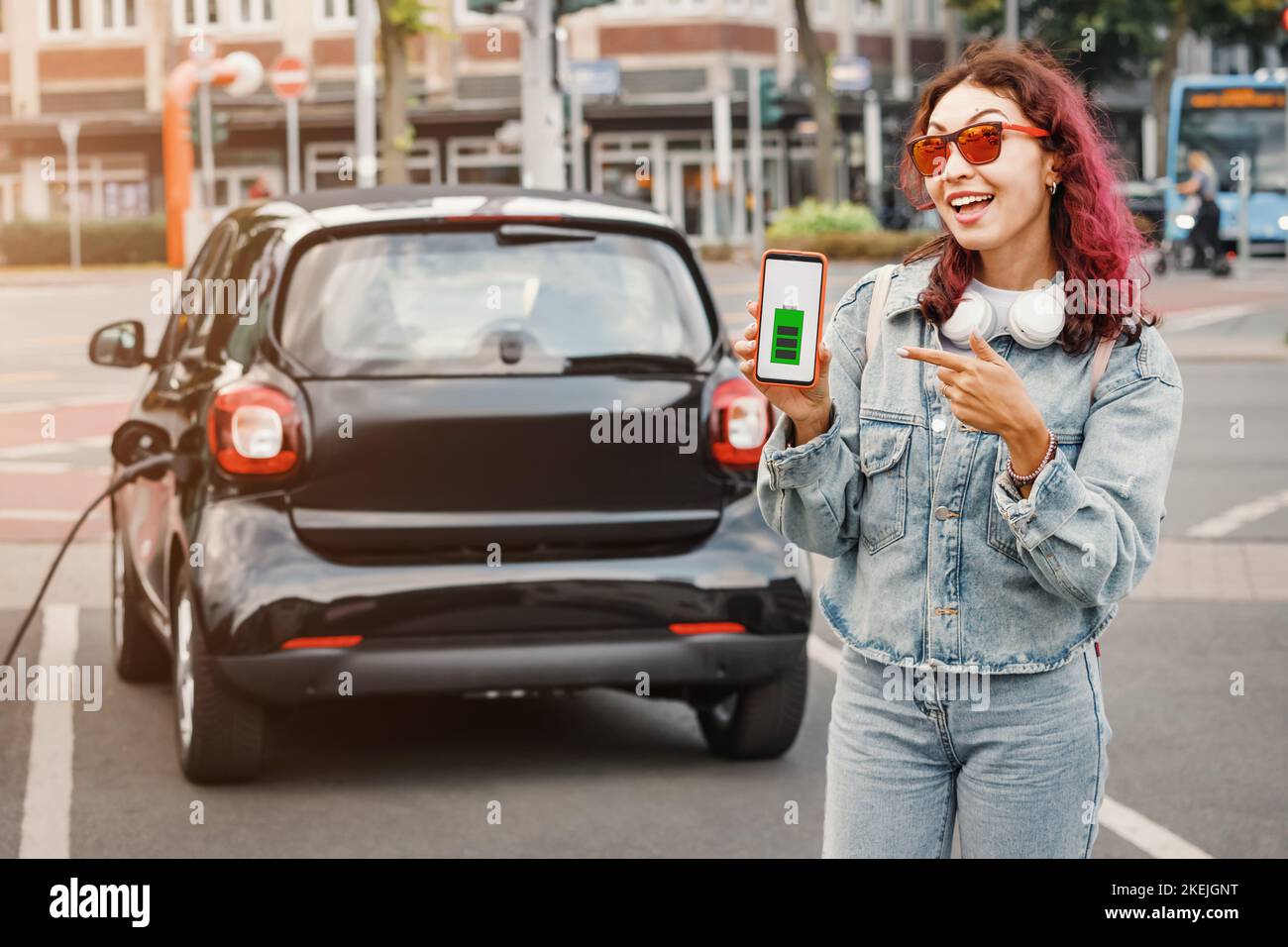 Girl shows the screen of a smartphone with a mobile application against the background of an electric car charging. Carsharing and eco transport Stock Photo