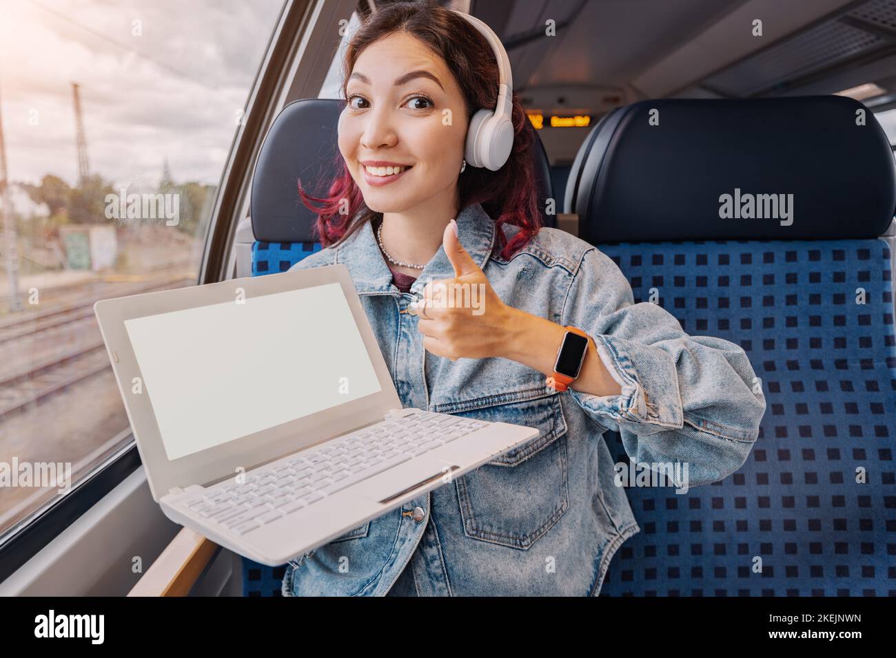 Happy young girl rides in a modern intercity train and shows white screen of her laptop. Work remotely via Wi-Fi Internet or study or watch a movie Stock Photo