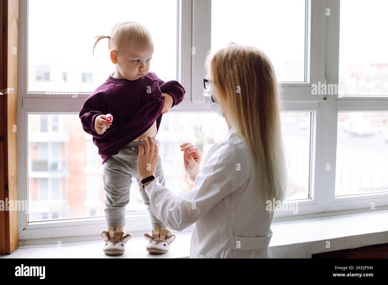 Worried little girl standing on windowsill, raising sweatshirt to show belly to childrens doctor wearing white uniform. Stock Photo