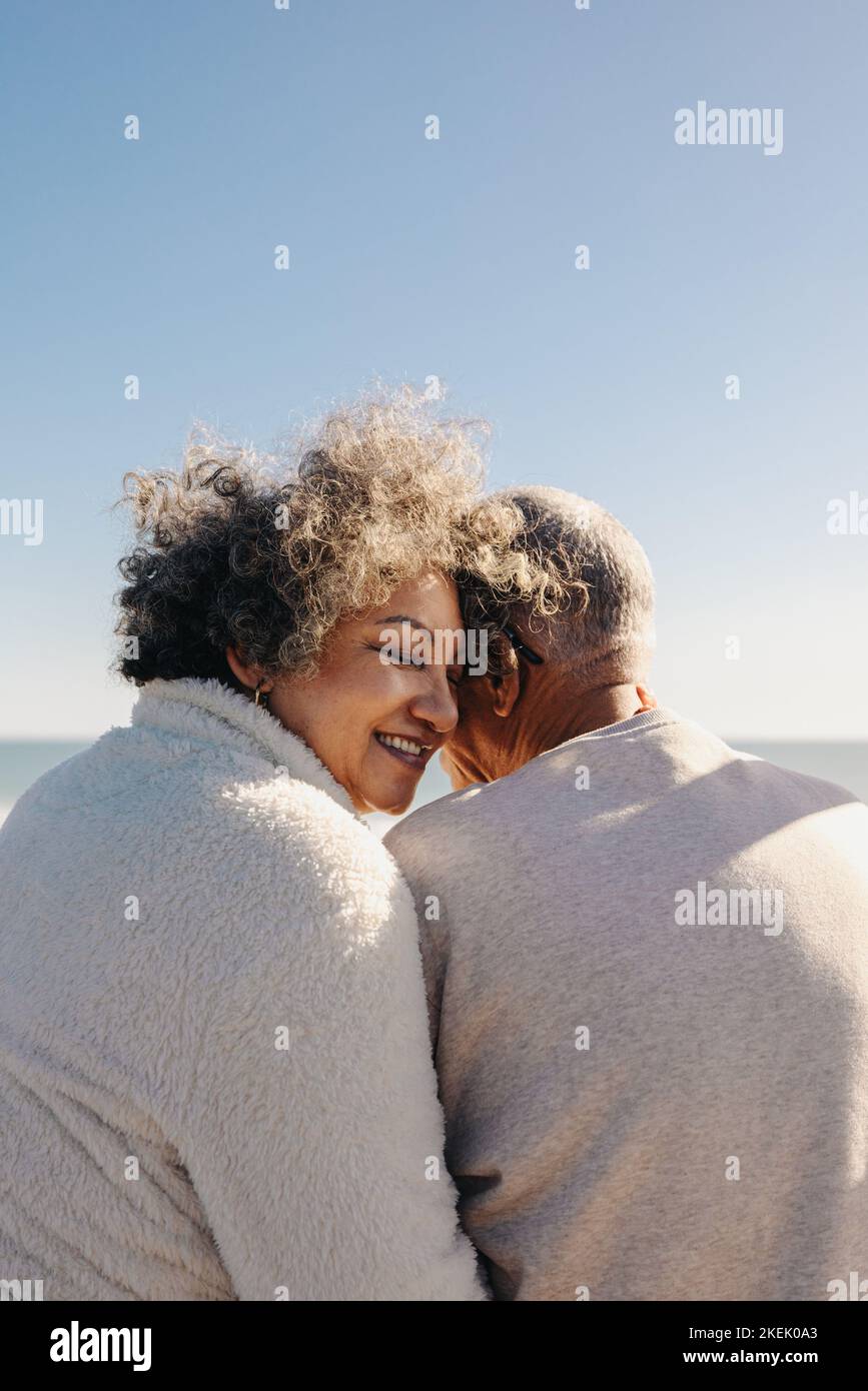 Back view of a happy senior woman smiling while sitting in front of the ocean with her husband. Retired elderly couple spending some quality time toge Stock Photo