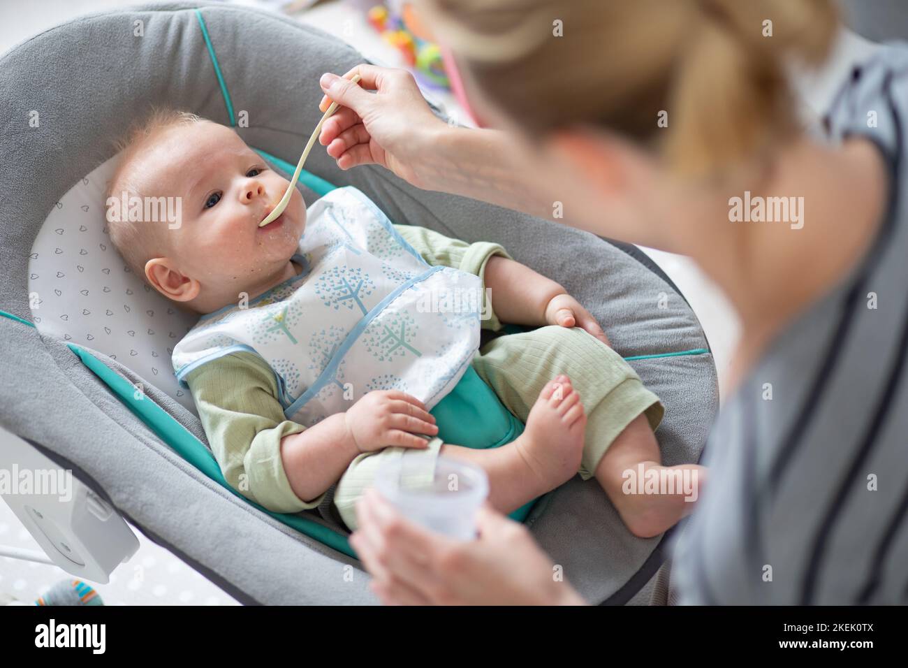 Mother spoon feeding her baby boy child in baby chair with fruit puree. Baby solid food introduction concept Stock Photo