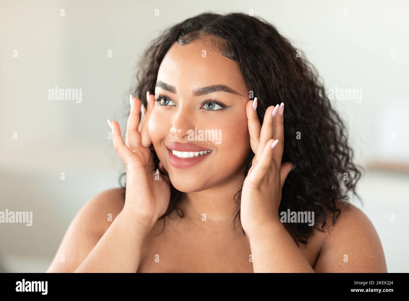 Beautiful black chubby lady touching her face and smiling while making beauty treatments at home, enjoying selfcare Stock Photo
