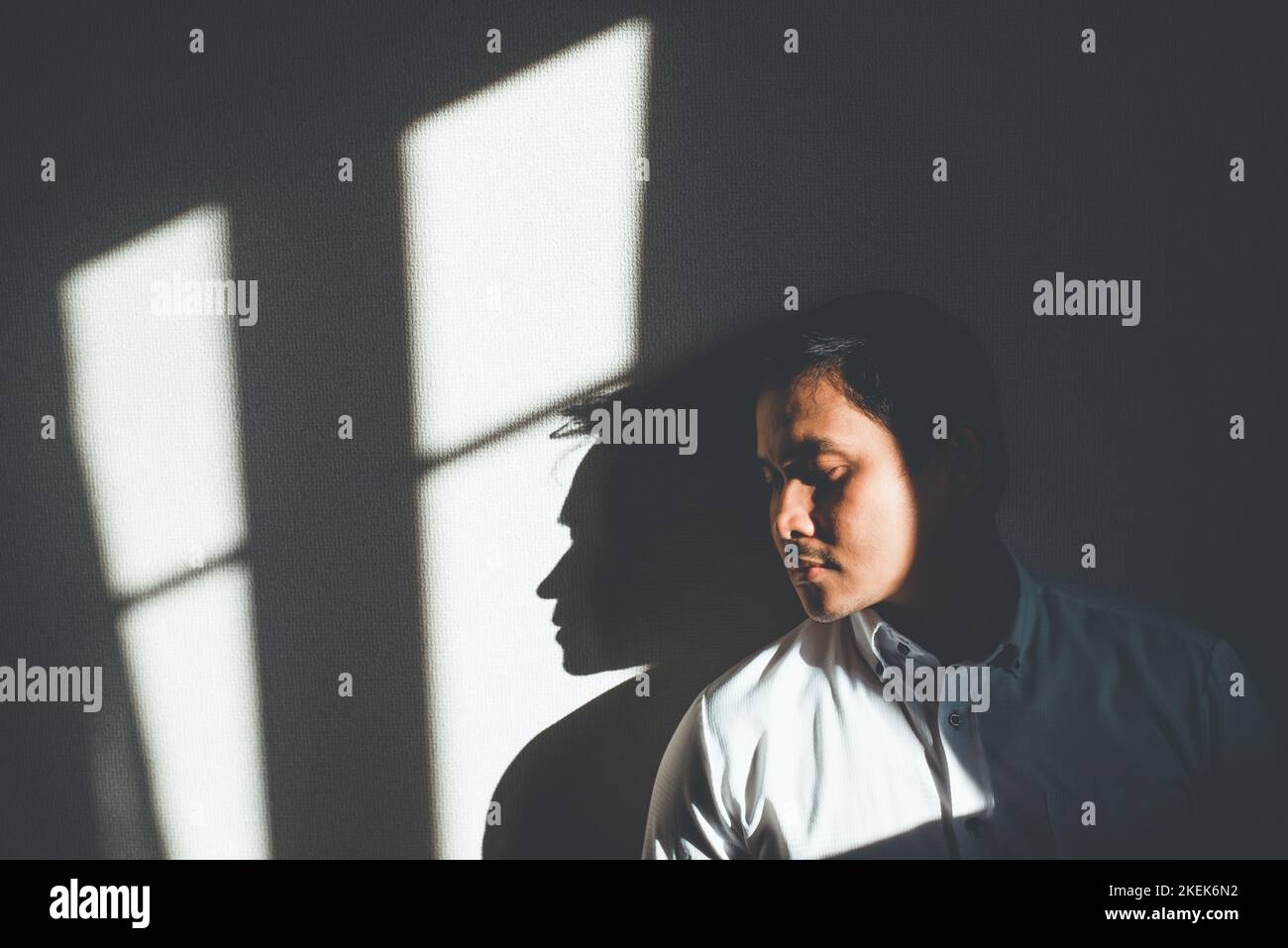 Depression. An depressed man leaning against the wall hit by a sunlight in an empty room. Stock Photo