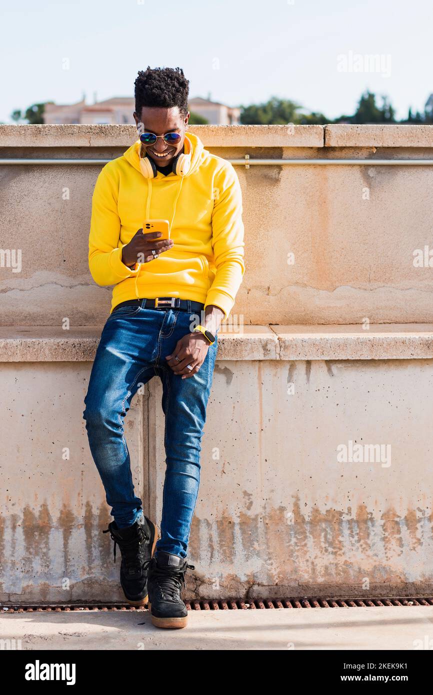 A smiling young black man dressed in a yellow sweatshirt, blue jeans and sunglasses is wearing while standing leaning against a wall. He is wearing he Stock Photo