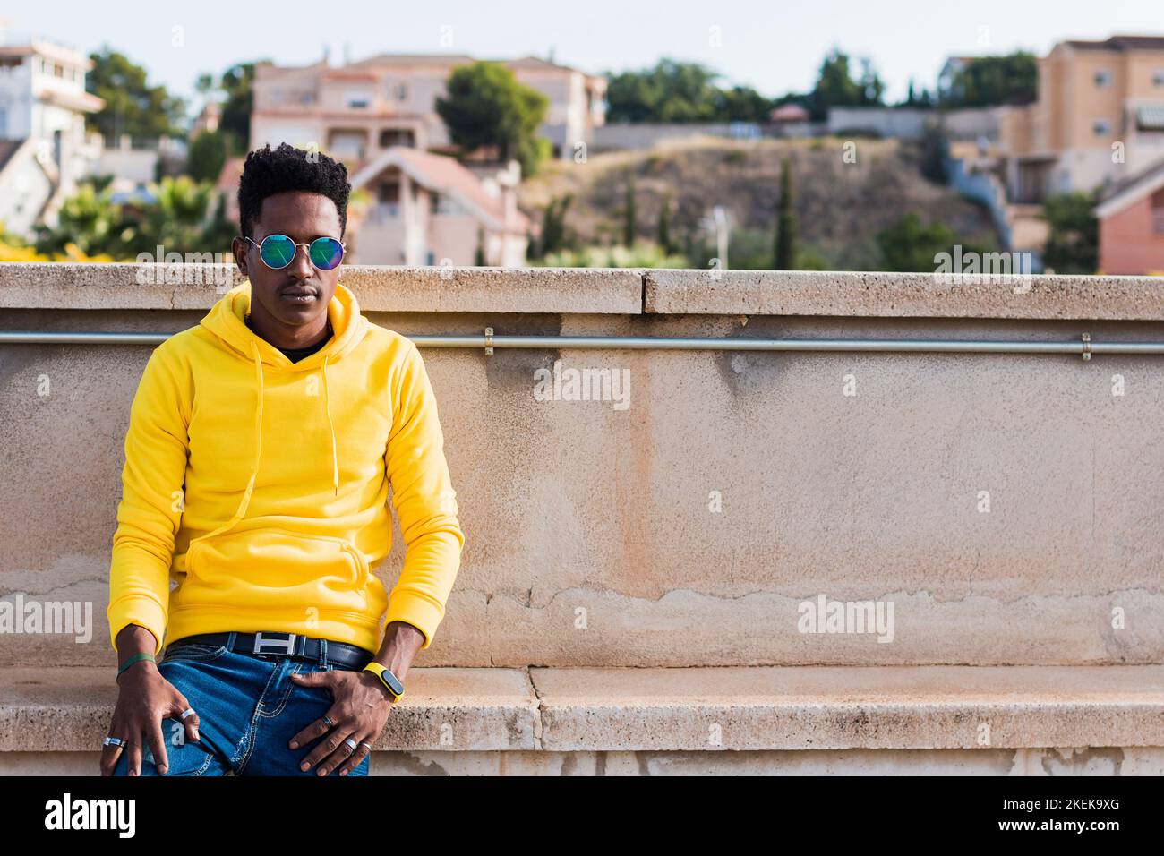 Horizontal plane portrait of a young black man wearing a yellow sweatshirt, blue jeans and glasses leaning against a low wall in the street. Out of fo Stock Photo