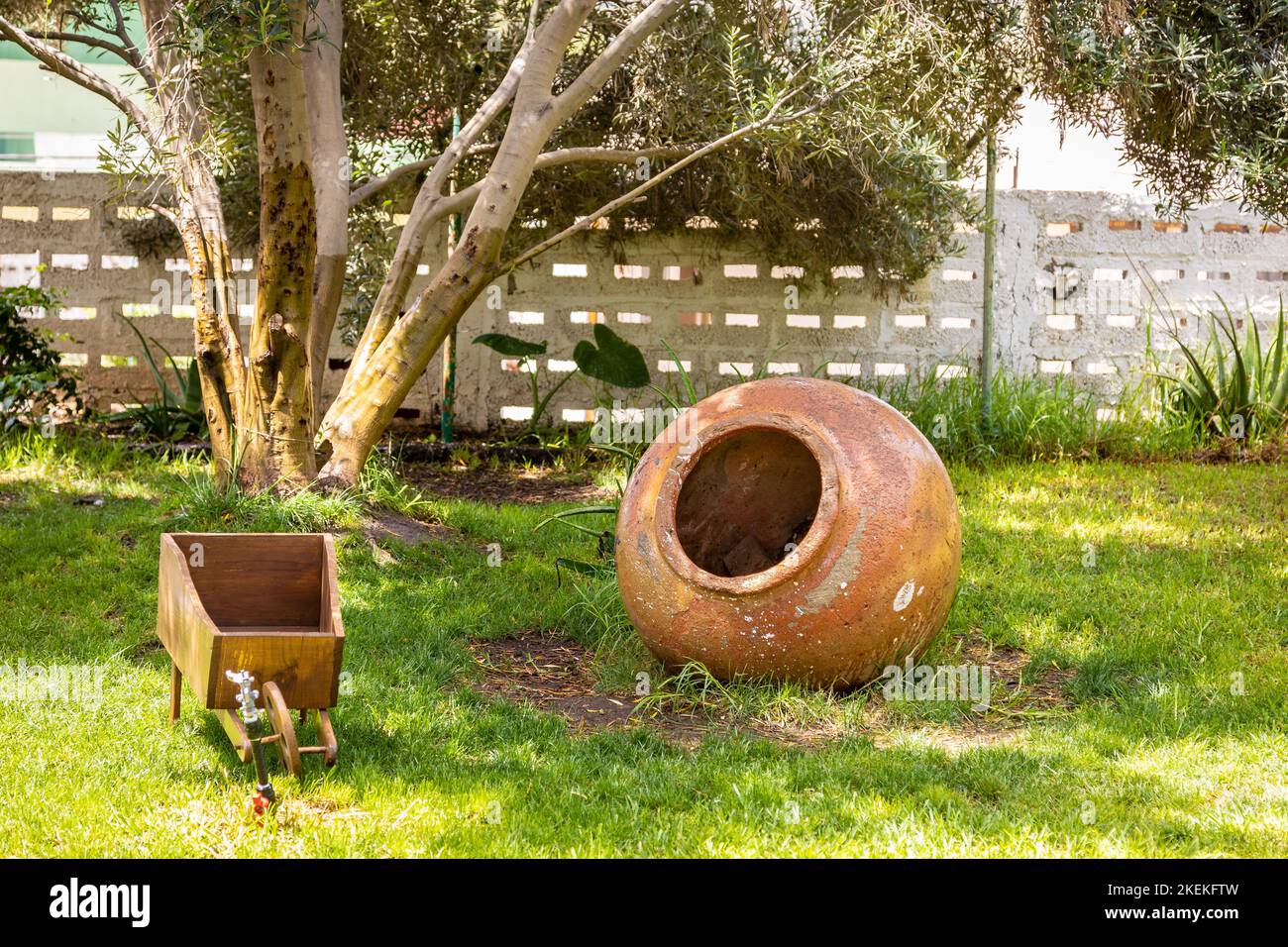 A shot of an old clay wine jug and a wooden wheelbarrow on the grass. Stock Photo
