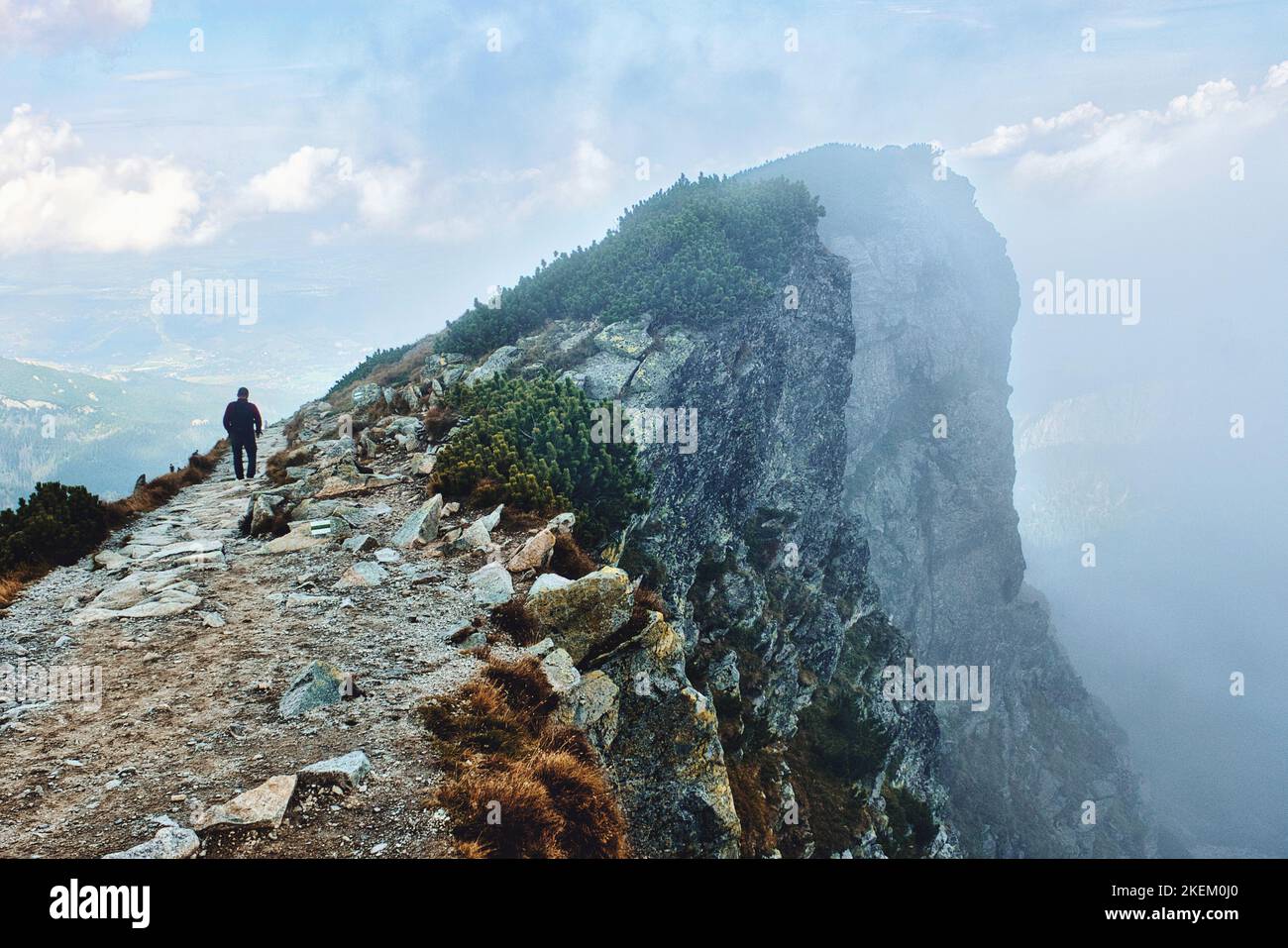 Lone hiker walking rocky mountain trail with steep rocky cliffs nearby covered in heavy fog and Poland, Zakopane town in distance in Tatra mountains Stock Photo