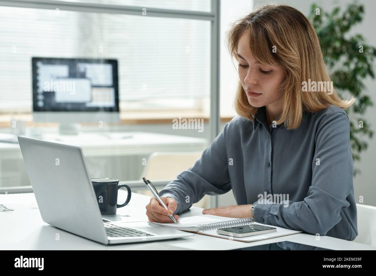 Young manager making notes in notepad while sitting at her workplace in front of laptop, she working at office Stock Photo