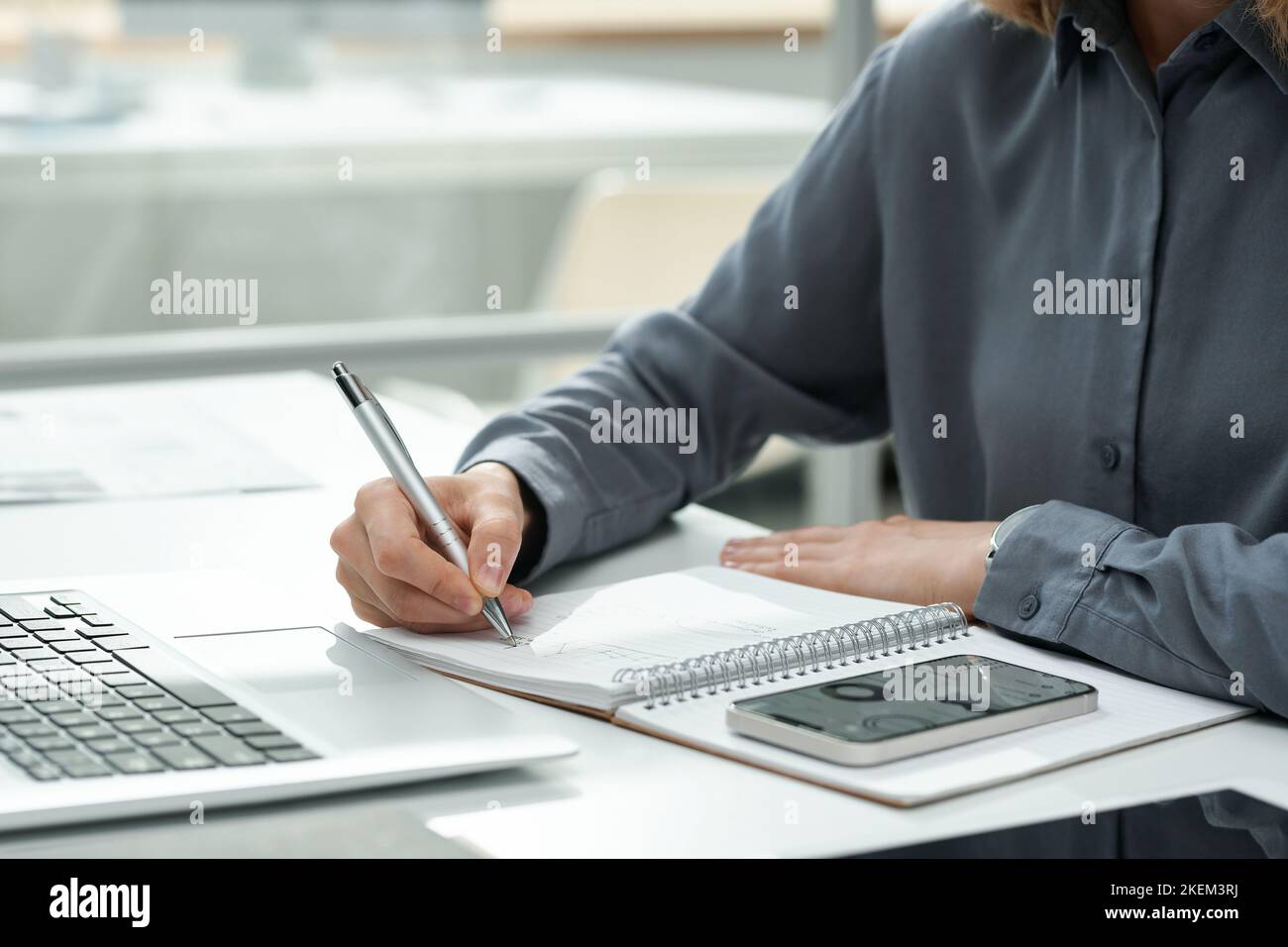 Close-up of young woman using smartphone with charts at her work while making notes in notepad at table Stock Photo