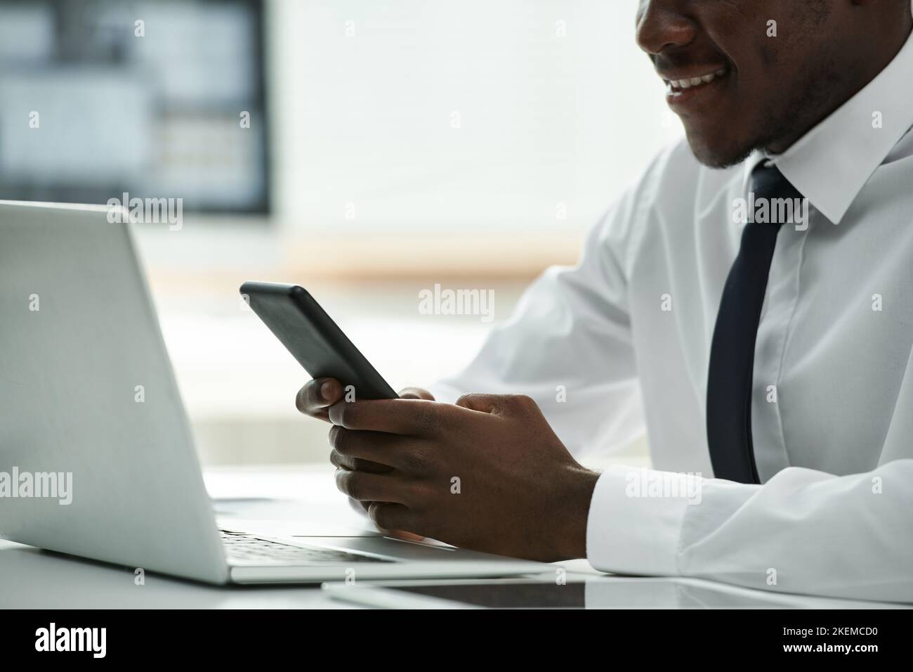 Close-up of African American businessman using smartphone and smiling while sitting at table in front of laptop Stock Photo
