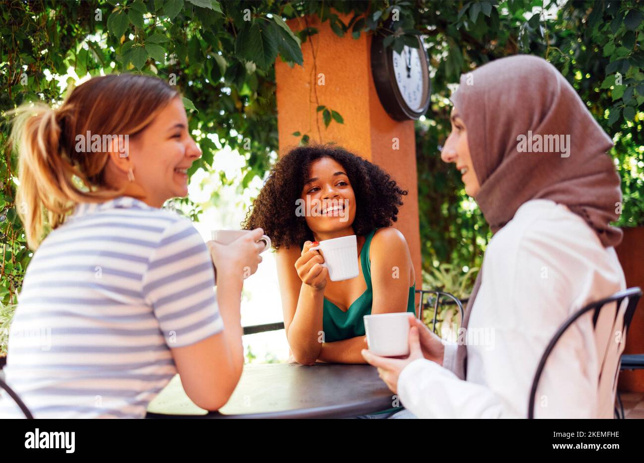 Multiethnic group of girls wearing casual clothes and traditional hijab bonding and having fun outdoors. Three young teen girls in garden cafe drinkin Stock Photo