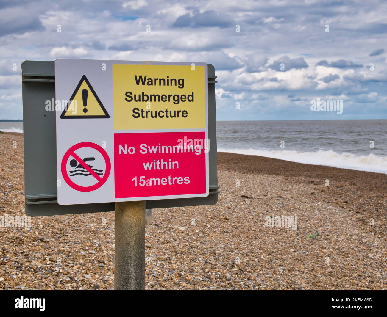 A red and yellow warning sign on a shingle beach at The Sluice on the Suffolk coast warns of a submerged structure and that swimming is not allowed. Stock Photo