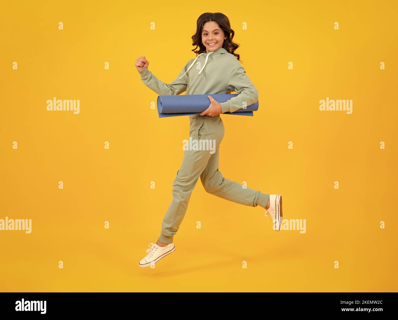Sportswear advertising concept. Run and jump. Teenager child girl in tracksuits jogging suit posing in the studio hold fitness mat. Stock Photo