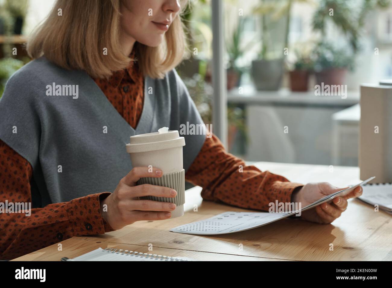 Young businesswoman drinking coffee from disposable cup while sitting at table and reading document Stock Photo