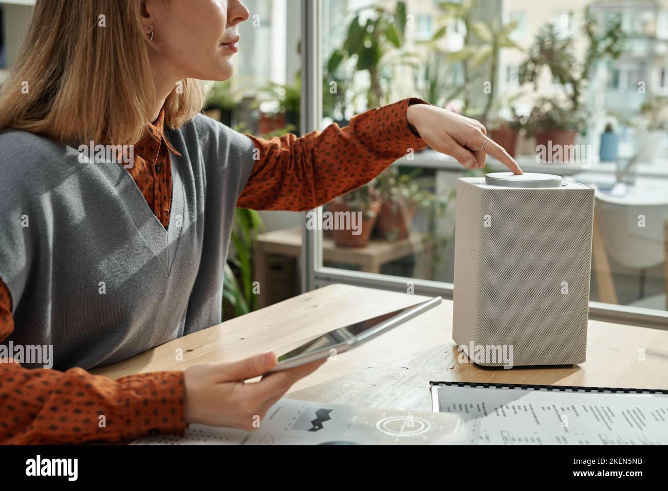 Young businesswoman turning on smart speaker and talking to it while working with tablet pc at table at office Stock Photo