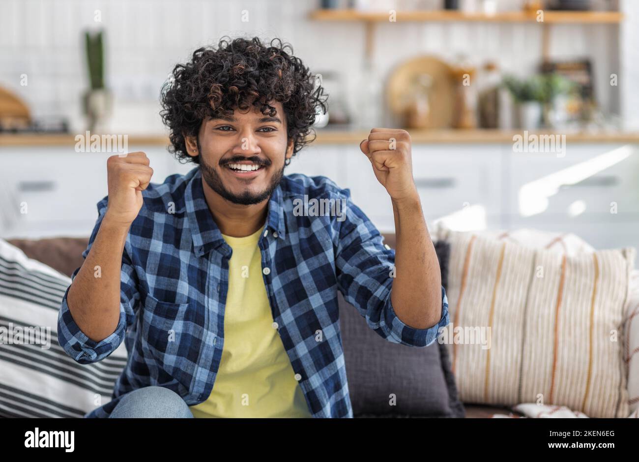 Smiling male with curly hair showing yes gesture, sitting on couch at home. Good news concept Stock Photo