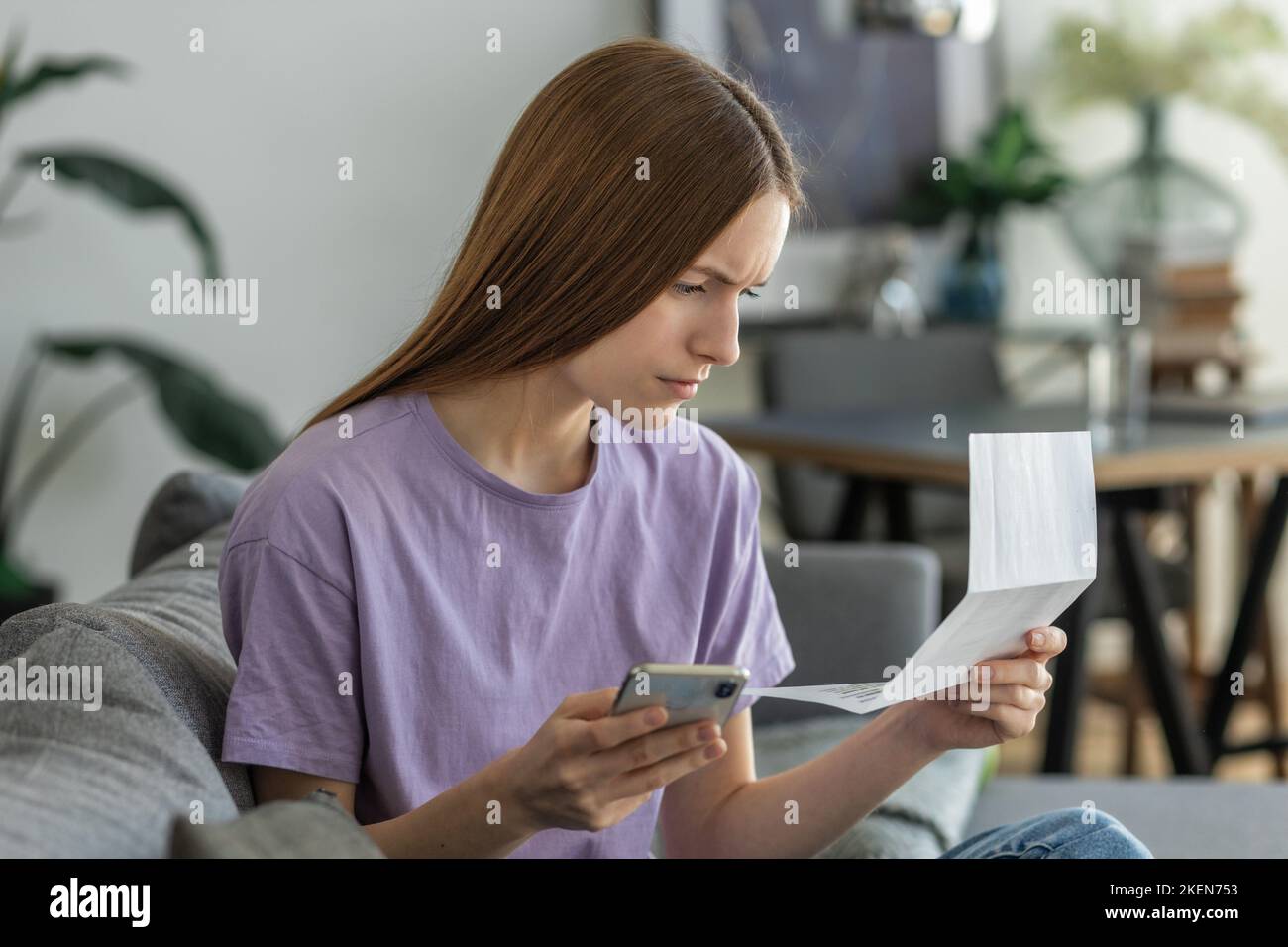 Worried young woman reading paper bill pay online using mobile phone at home. Female holding bank letter reading Stock Photo