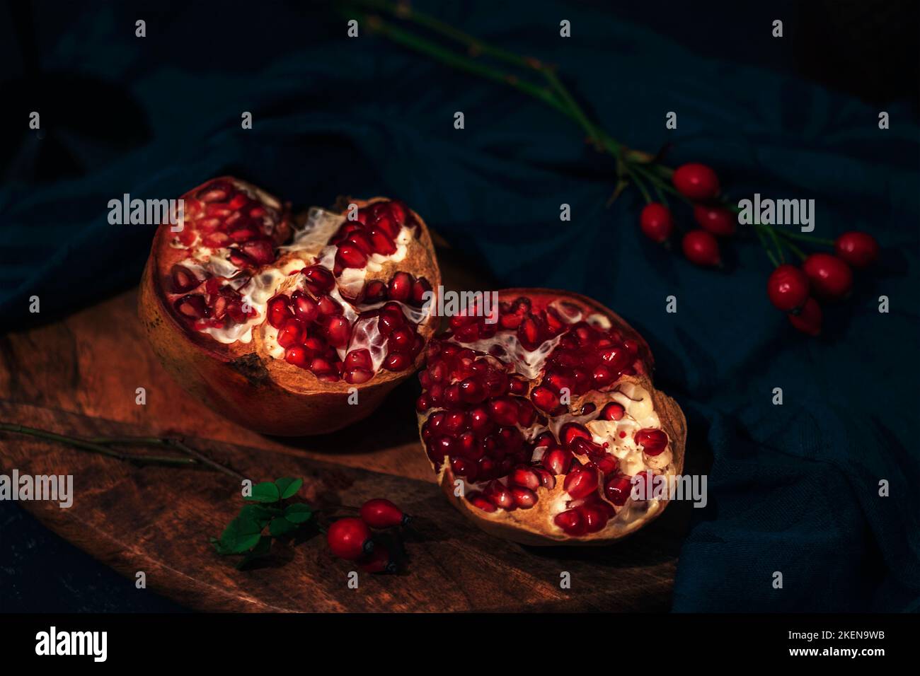 Pomegranate on a cutting board and rosehip branches on black table. Still life. Closeup. Stock Photo