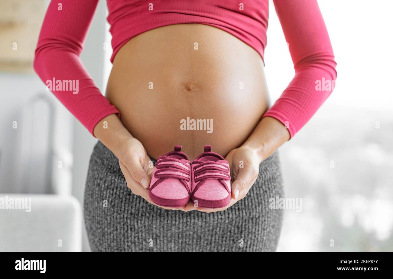 Pregnancy. Pregnant woman expecting baby girl holding pink red shoes in gender reveal. Expecting mom showing baby bump belly. Baby clothing Stock Photo