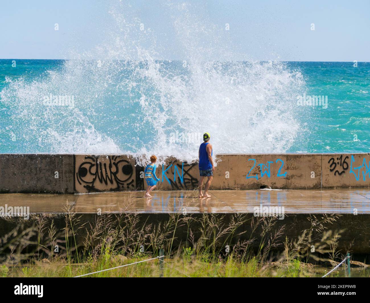 A father and his young son brave the seawall as a large wave crashes over it on a windy summer day. Montrose Point, Chicago, Illinois. Stock Photo