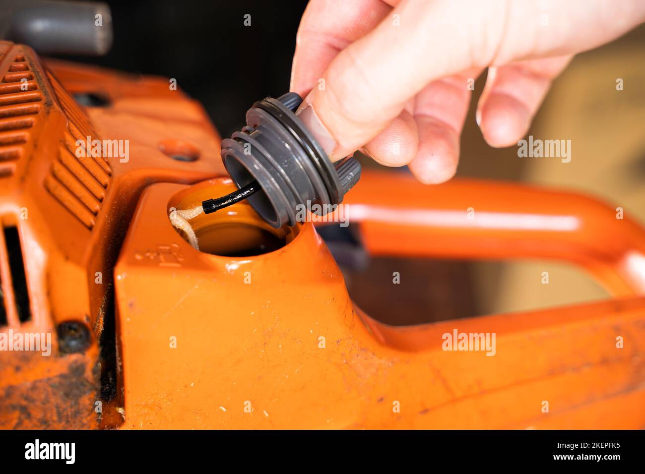 A hand unscrews the cap of the fuel mixture tank in an orange chainsaw