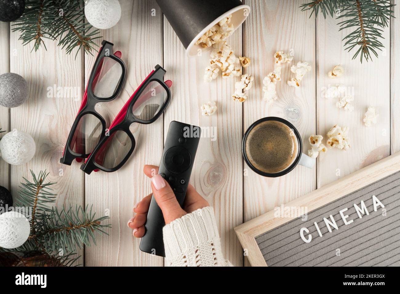 Female hand hold TV remote controller, top view. Watching movie film at home with pair of cinema glasses, cup of coffee and popcorn. Spruce branches Stock Photo