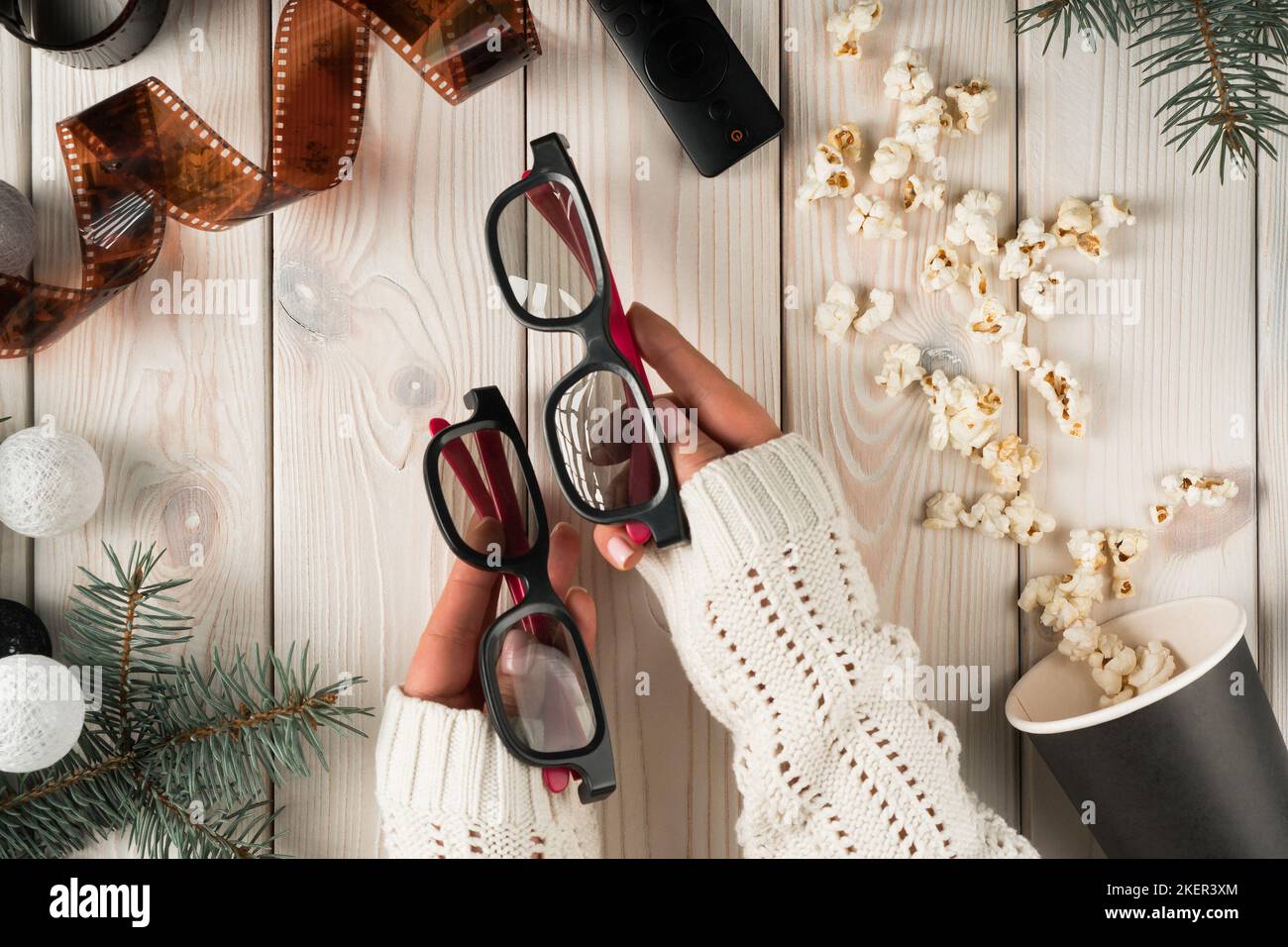 Female hands hold pair of cinema glasses for watching movie film at home, top view. Decorative spruce branches and christmas tree balls. Eating Stock Photo