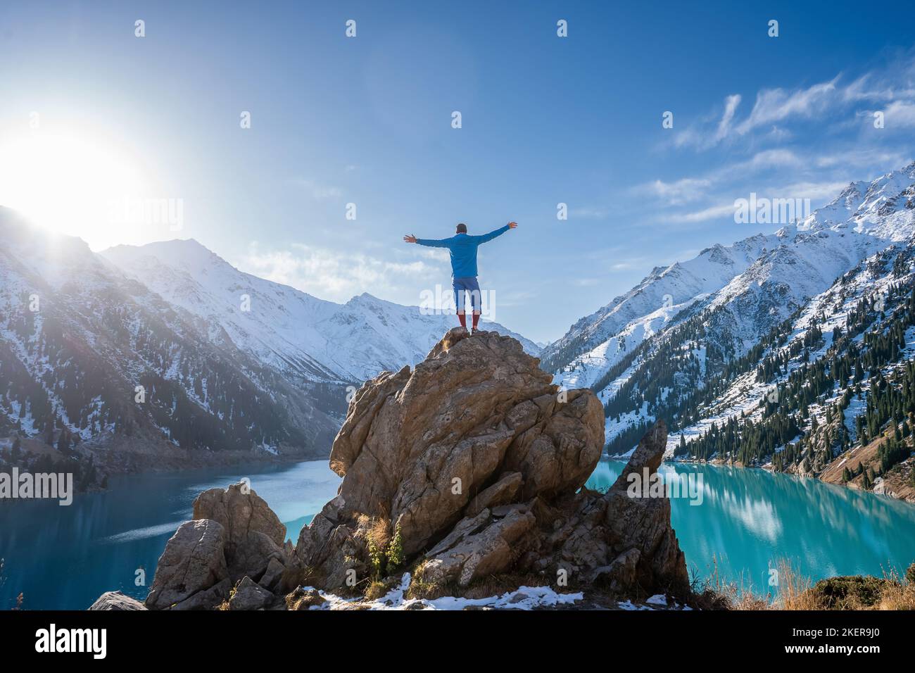 Longshot of a man standing on a huge boulder on a vantage point over stunning turquoise lake surrounded by snowy mountains on a sunny day. His hands a Stock Photo