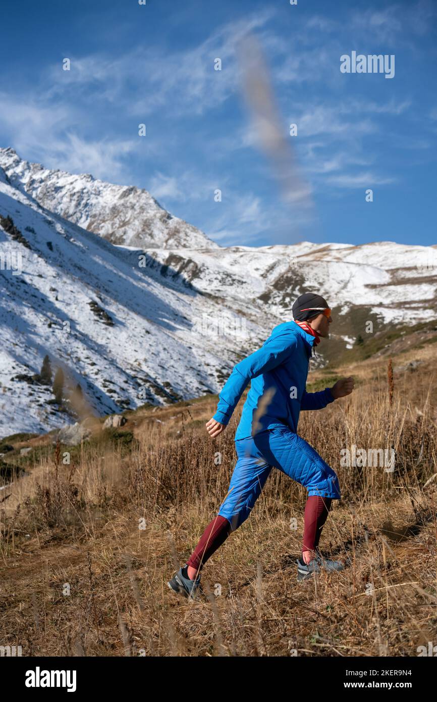 Dressed up man in sportswear hiking a grassy slope. In warm wind resistant clothes. Next to a snowy mountain. Stock Photo