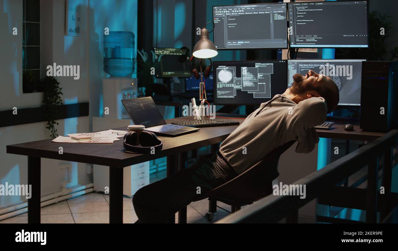 Male developer enjoying programming success at night, working on coding new user interface. Feeling cheerful and pleased about server system developing, using multiple monitors. Handheld shot. Stock Photo