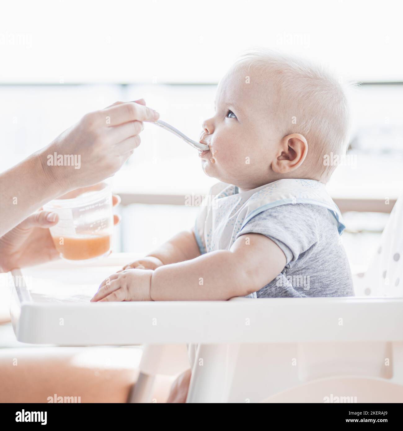 Mother spoon feeding her baby boy child in baby chair with fruit puree on a porch on summer vacations. Baby solid food introduction concept Stock Photo