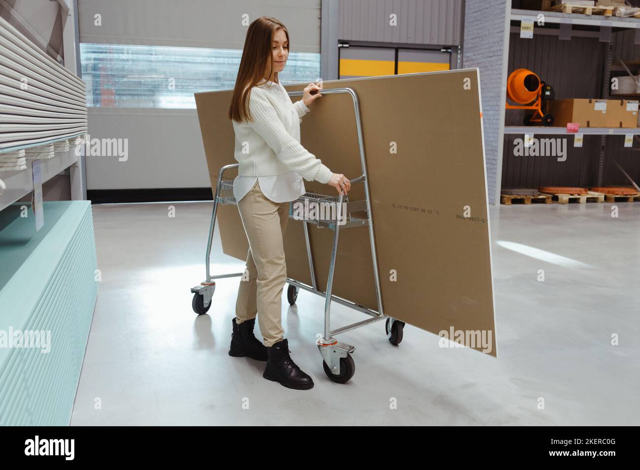 Young woman carrying trolley cart with drywall sheets in hardware store