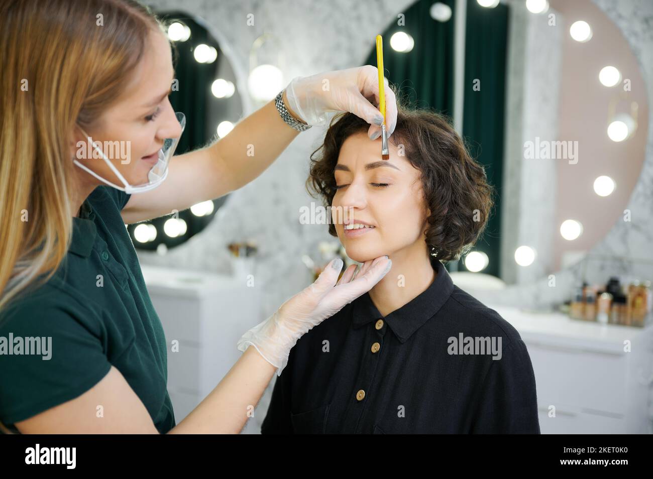 Happy woman keeping eyes closed and smiling while having eyebrow correction procedure in beauty salon. Female beauty specialist in sterile gloves shaping client eyebrows with cosmetic brow brush. Stock Photo