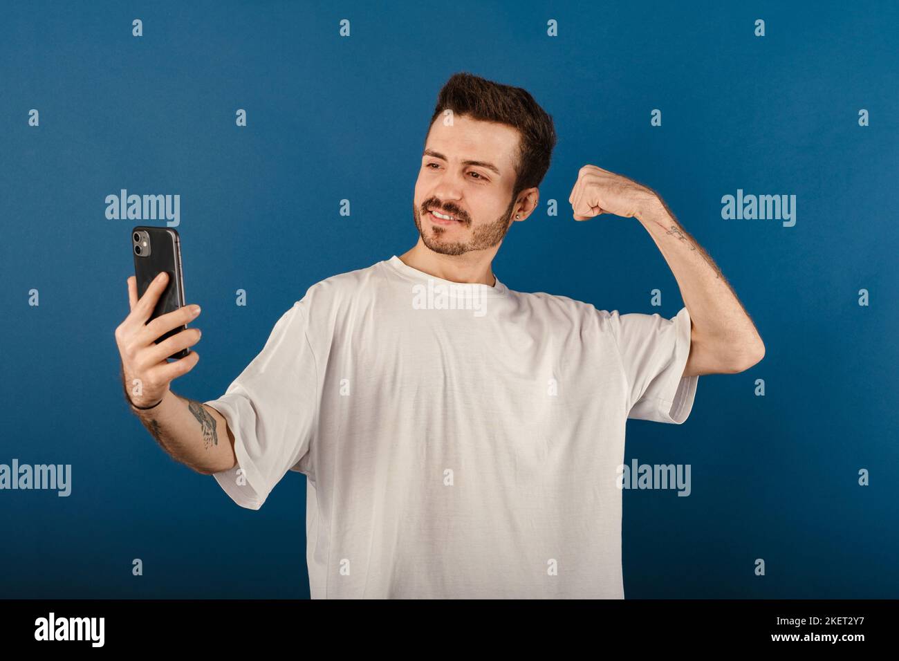 Portrait of cheerful man wearing white t-shirt posing isolated over blue background taking selfie, showing strong arm, flexing biceps and posing for p Stock Photo