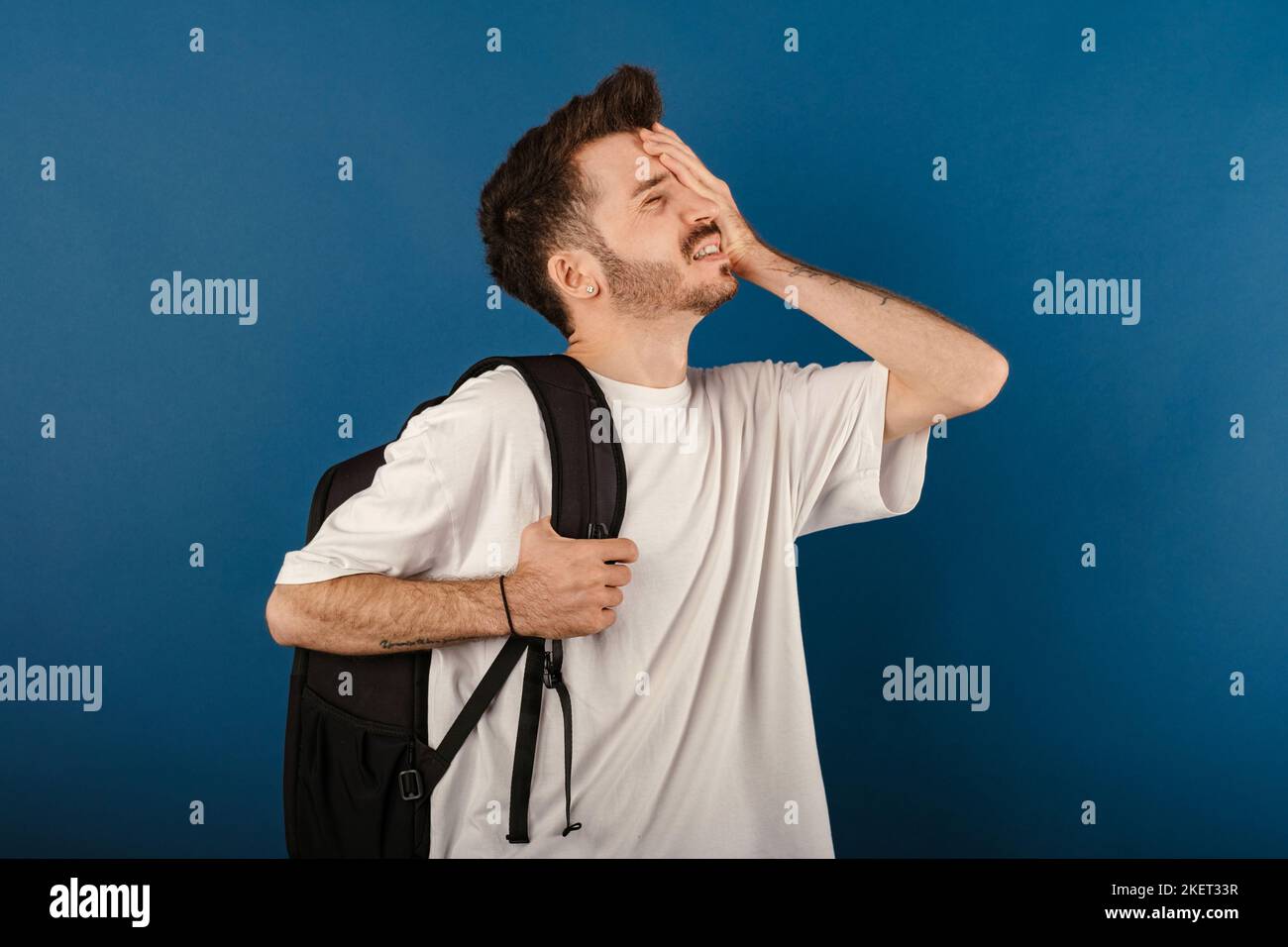 Caucasian young man wearing t-shirt posing isolated over blue background suffering headache and holding hands on forehead. Education in university col Stock Photo