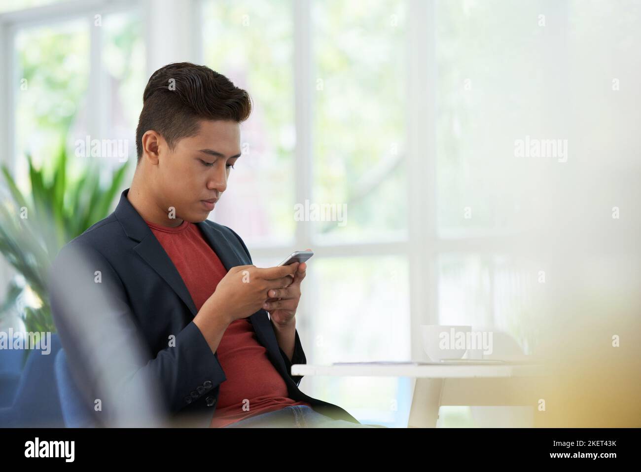 Confident young man sitting at cafe table with cup of coffee and browsing Internet on modern smartphone, panoramic windows on background Stock Photo