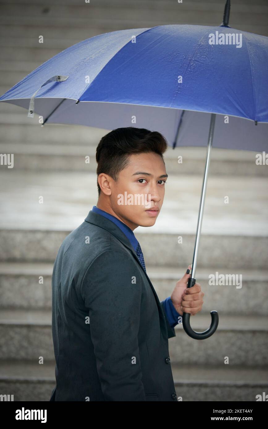 Waist-up portrait of confident Vietnamese entrepreneur wearing elegant suit looking away while walking upstairs with umbrella in hand Stock Photo