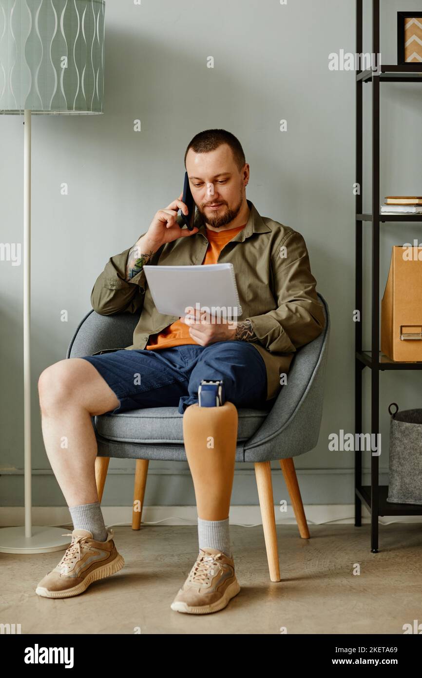 Full length portrait of man with prosthetic leg calling by phone while sitting in chair in home interior Stock Photo
