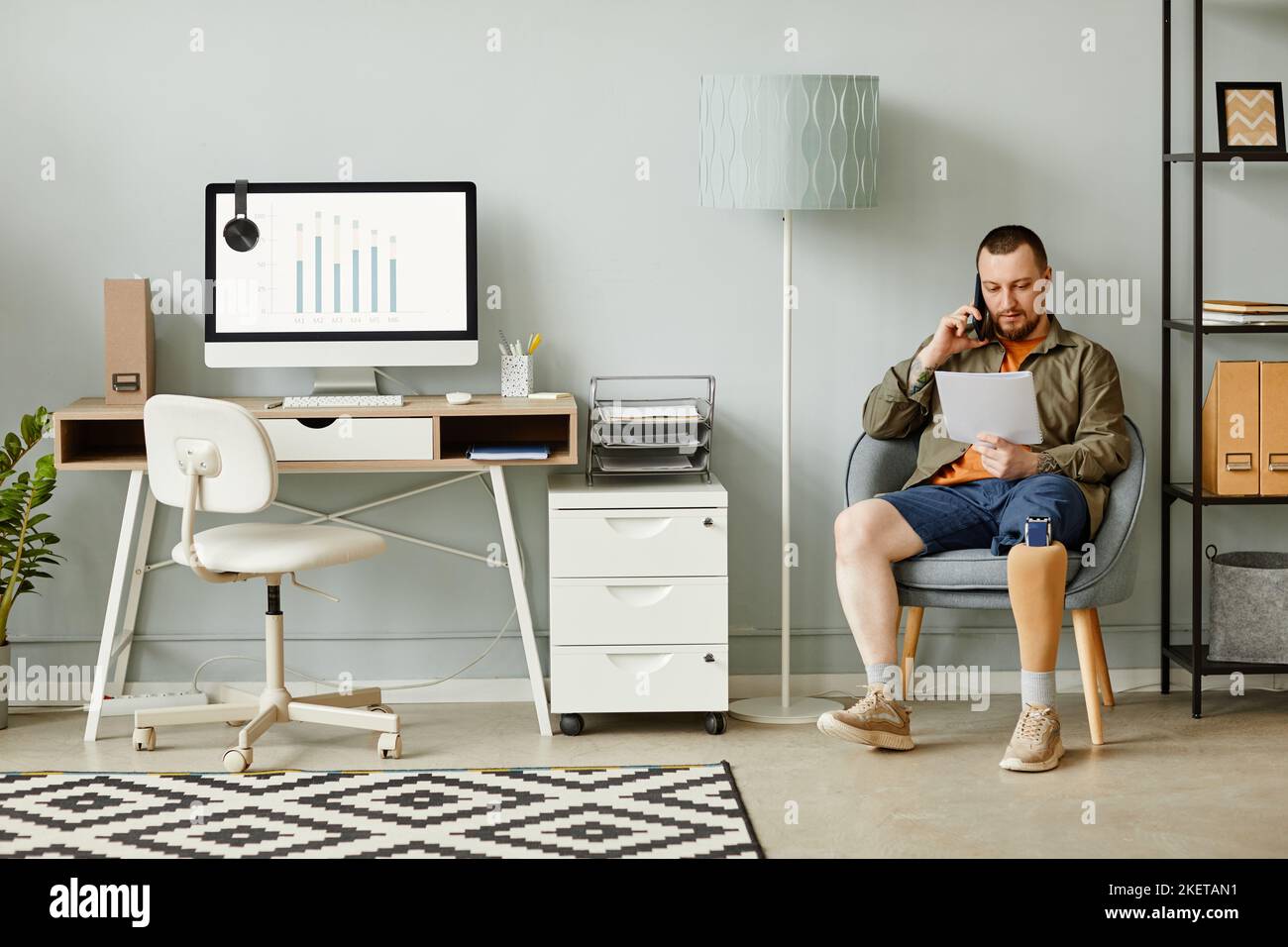 Full length portrait of man with prosthetic leg calling by phone while sitting in chair in home office interior, copy space Stock Photo