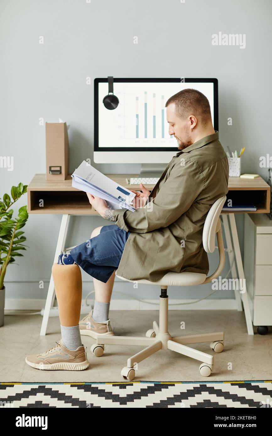 Vertical full length portrait of man with prosthetic leg working at home office and reading documents Stock Photo