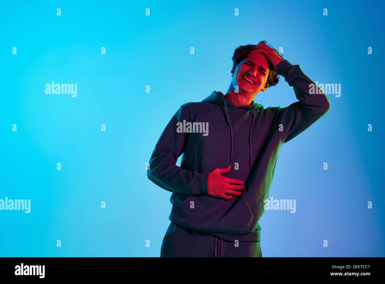 Portrait of young man with curly hair posing, emotively laughing isolated over blue background in neon light. Extremely funny Stock Photo