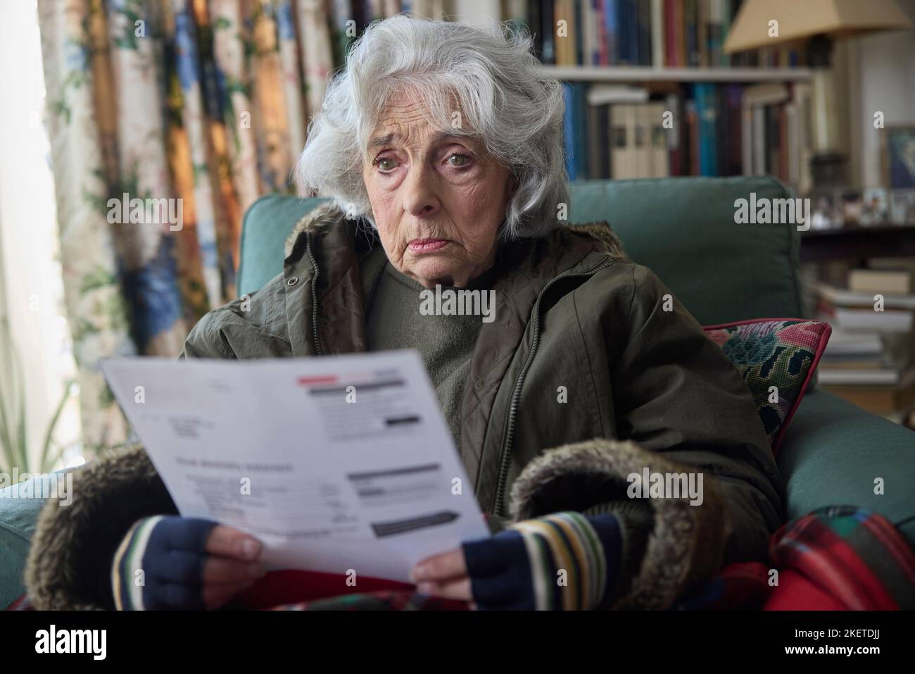Senior Woman Wearing Coat Indoors Trying To Keep Warm At Home In Energy Crisis Looking At Energy Bill Stock Photo