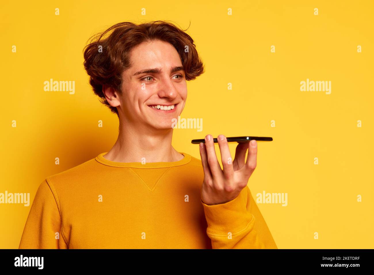 Portrait of young man with curly hair posing, recording voice message on phone isolated over yellow background Stock Photo