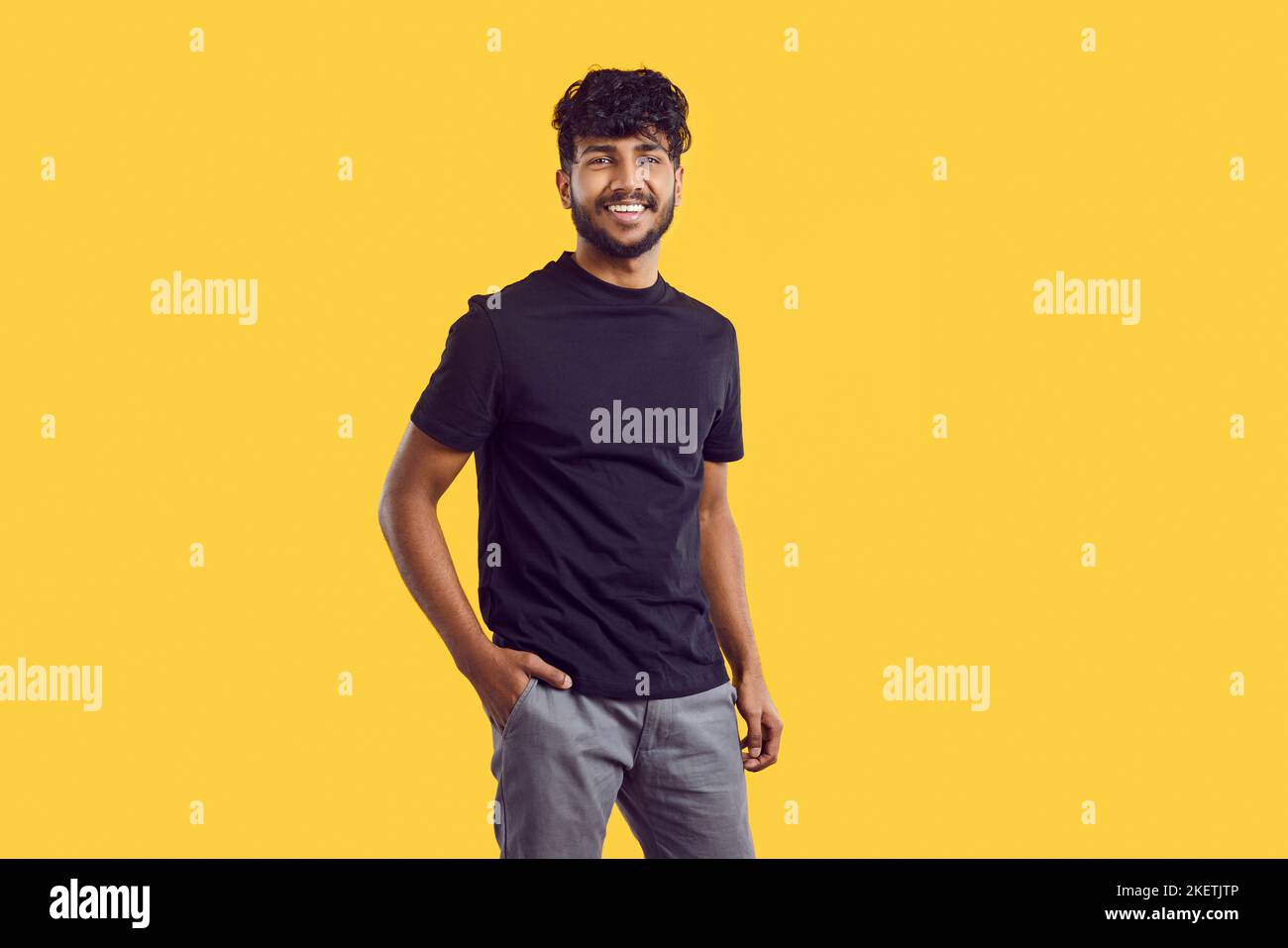 Young South Asian man with dark curly hair posing with smile in casual clothes stands in studio Stock Photo